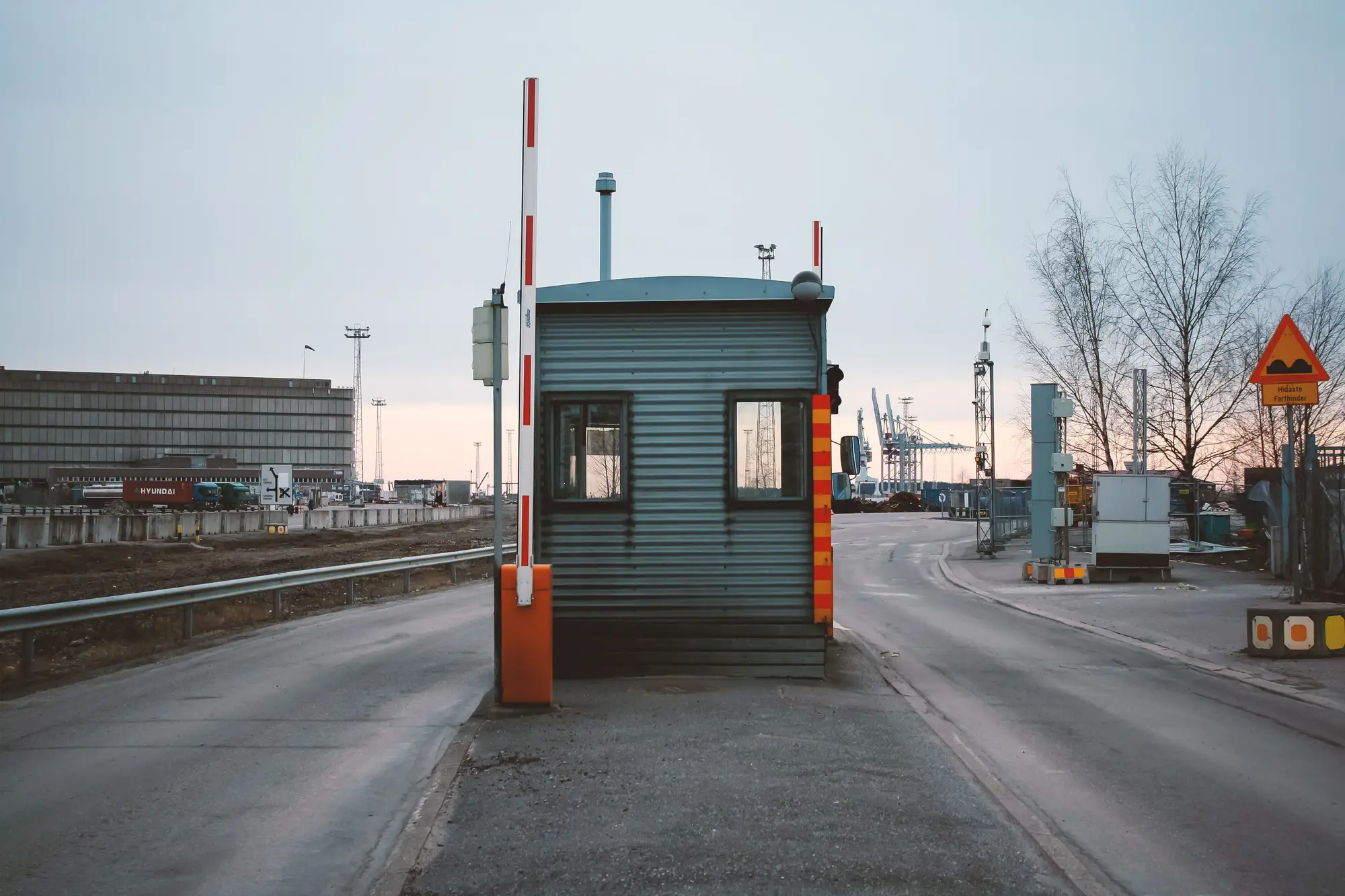 A gatehouse and striped barrier on an empty approach road at a port terminal, cranes in the distance